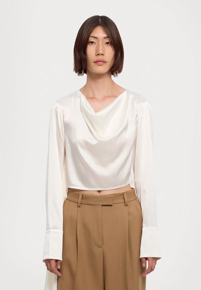 Κομμένο κρεμ σατέν τοπ με κρεμαστή λαιμόκοψη και μακριά μανίκια. Συνδυασμένο με καφέ παντελόνι υπερύψωσης, ραμμένο με πιέτες.