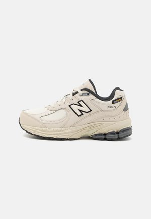 Baskets de course beige avec tige en maille et synthétique, accents noirs, col rembourré et semelle amortie. Présente le logo "2002R" et une étiquette de durabilité.