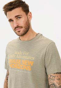 Lys grøn t-shirt med korte ærmer, rund halsudskæring og trykt orange tekst, der står "Skabt til daglige eventyr JAGT NYE HORIZONTER."