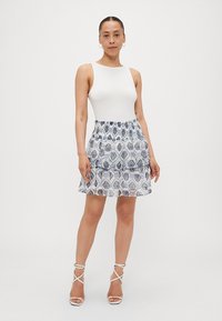 Top blanco sin mangas con una falda estampada, que presenta volantes de capas en tonos azul y blanco, y tacones blancos con tiras.