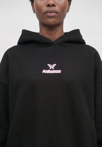 Person trägt einen schwarzen Hoodie mit einem pinken Schmetterling und dem Wort "Felicious" auf der Brust, gezeigt vom Kinn bis zur Mitte des Oberkörpers.