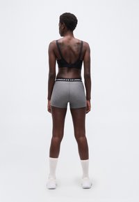 Γυναίκα που στέκεται με την πλάτη στραμμέ νη, φορώντας μαύρο αθλητικό σουτιέν και γκρι αθλητικό σορτς Under Armour, λευκές κάλτσες και λευκά αθλητικά παπούτσια.