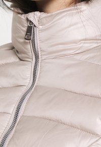 Padded jacket w miękkim beżowym materiale z wysokim kołnierzem. Posiada srebrny suwak i pikowaną fakturę, zapewniającą ciepło i izolację.