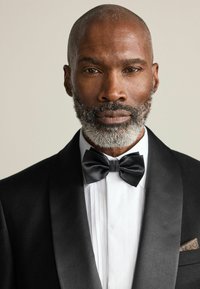 Black tuxedo jacket with satin lapels, white pleated shirt, and black bow tie. Grey beard and short hair, with a patterned pocket square.