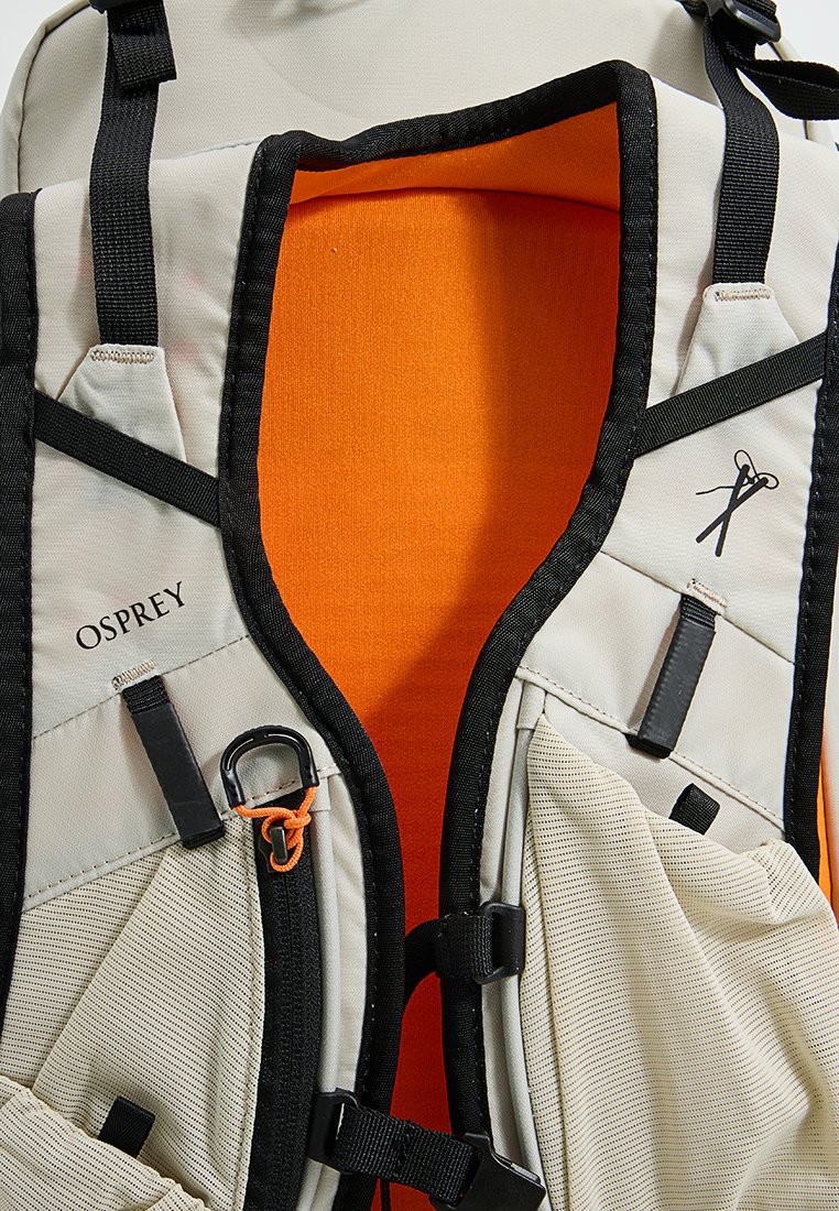 Chaleco de senderismo beige con detalles en negro, correas ajustables, bolsillos de malla y un forro interior naranja; presenta el logo de Osprey y herrajes.