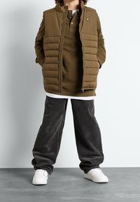 Gilet imbottito verde oliva con zip, indossato sopra un polo a maniche lunghe marrone. Abbinato a pantaloni in velluto a coste grigi scuri e sneakers bianche.