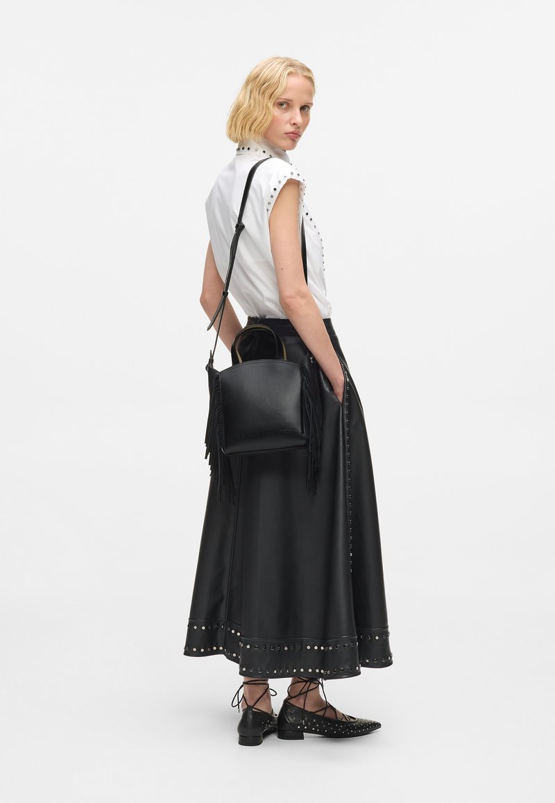 Fekete bőr, bojtokkal díszített kézi táska, állítható pánttal, amit egy fényes fehér inggel párosítanak, amely szegecsekkel van díszítve, és egy fekete bőr szoknyával.