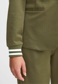 Veste vert olive avec poignets côtelés arborant des rayures blanches et vert foncé. Tissu doux, coupe ajustée, avec une poche unique sur le côté gauche.