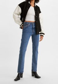 Chaqueta varsity en blanco y negro con un corte relajado, combinada con jeans de tiro alto azules y botines negros de tacón. Texturas suaves visibles.