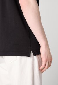 Sort bomulds T-shirt med en sideslids i kanten, parret med hvide denimbukser. Nærbillede af skjortens tekstur og pasform.
