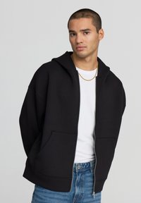 Giovane uomo che indossa una felpa nera con zip, una maglietta bianca, una collana in oro e jeans blu, in piedi contro uno sfondo grigio uniforme.