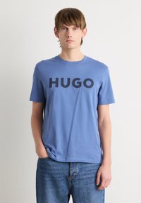 Mėlynas medvilnės marškinėliai su dideliu juodu "HUGO" logotipu priekyje. Apvalus kaklas, trumpomis rankovėmis, laisvas siluetas ir švelni tekstūra.
