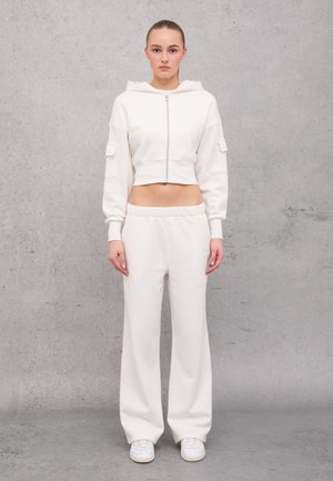 Felpa cropped color crema con chiusura a zip e due tasche laterali, abbinata a pantaloni della tuta a gamba larga coordinati. Tessuto morbido e accogliente.