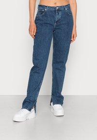 Jeans de ganga azul com um design de perna reta, leve textura desbotada e aberturas laterais com fecho, combinados com ténis plataforma brancos.