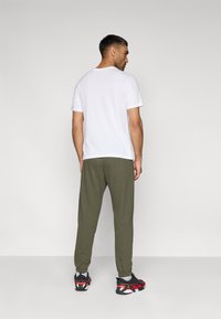 Λευκό κοντομάνικο μπλουζάκι, χακί πράσινο παντελόνι φόρμας, μαύρα και κόκκινα αθλητικά παπούτσια, στέκεται με την πλάτη προς τα εμπρός σε απλό φόντο.