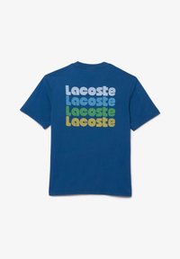 Camiseta de algodón azul con un diseño en la parte posterior que presenta el texto repetido "Lacoste" en colores variados: azul claro, verde y amarillo. Mangas cortas.