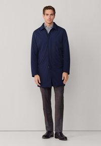 Man staat in een marineblauwe jas over een gestreept shirt, grijze broek en zwarte leren schoenen tegen een effen lichte achtergrond.