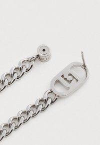 Sudraba metāla ķēdes aproce ar lielu ovālu gredzenu, kurā ir izgriezts logo, un apļu sprādzi ar iegravētu zīmola nosaukumu. Gluda, spīdīga virsma.