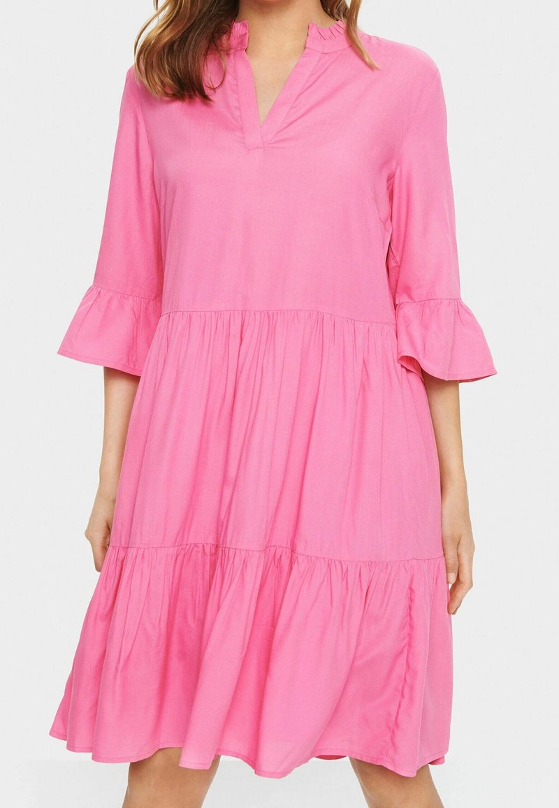 Femme portant une robe ample rose longue jusqu'au genou avec des manches cloche trois-quarts et un petit col froufroutant.