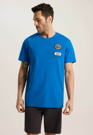 Niebieska bawełniana koszulka z okrągłym dekoltem, krótkimi rękawami i dwoma haftowanymi naszywkami na piersi. W połączeniu z ciemnymi szortami.