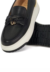 Black leather loafer with textured surface, featuring decorative laces and a metallic accent. Thick white sole with a patterned black tread.