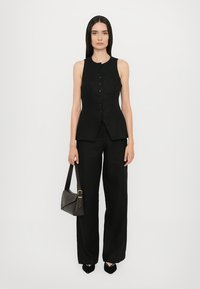 Black sleeveless vest with a buttoned front and pointed hem, paired with wide-leg black trousers. Model holds a dark handbag.