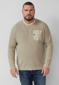 Beżowy sweter o splocie waflowym z okrągłym dekoltem, z kontrastową kieszenią. Dopasowane niebieskie dżinsy. Teksturowana tkanina i luźny krój.