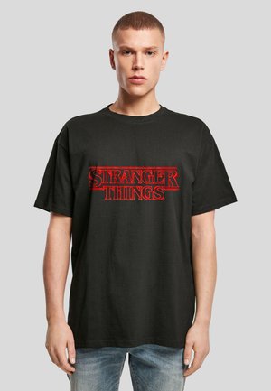 Bărbat tânăr cu păr scurt poartă un tricou negru supradimensionat cu text roșu „Stranger Things”, asortat cu blugi albaştri decolorați.