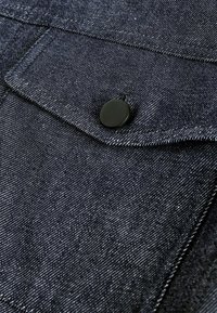 Tume denim kangas, millel on peen sära, ja silmapaistev tasku musta nööp/klõpsuga. Õmblused on nähtavad servade mööda.