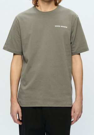 Person trägt ein einfarbiges olivgrünes kurzärmeliges T-Shirt mit weißem "WOOD WOOD" Text auf der oberen linken Brustseite und schwarze Hose.