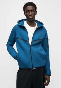 Nike tumši zaļš džemperis ar rāvējslēdzēju un melniem akcentiem, ar rāvējslēdzēja kabatu uz kreisās rokas, mīkstu audumu un brīvu pieguļošu stilu, kombinēts ar tumšām biksēm.