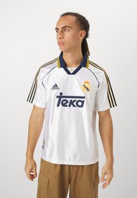 Maillot de football blanc du Real Madrid avec col marine, rayures jaunes et logo "Teka" noir. Manches courtes, fabriqué en matériau synthétique lisse.