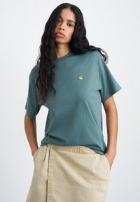 Τυρκουάζ βαμβακερό t-shirt με στρογγυλή λαιμόκοψη και κοντά μανίκια, με μικρό κίτρινο λογότυπο στη αριστερή πλευρά του στήθους. Απαλή, χαλαρή εφαρμογή.