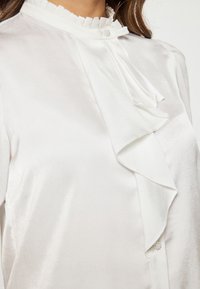 Blusa bianca in satin con colletto arruffato, pieghe centrali e bottone decorativo. Tessuto liscio con una leggera lucentezza.