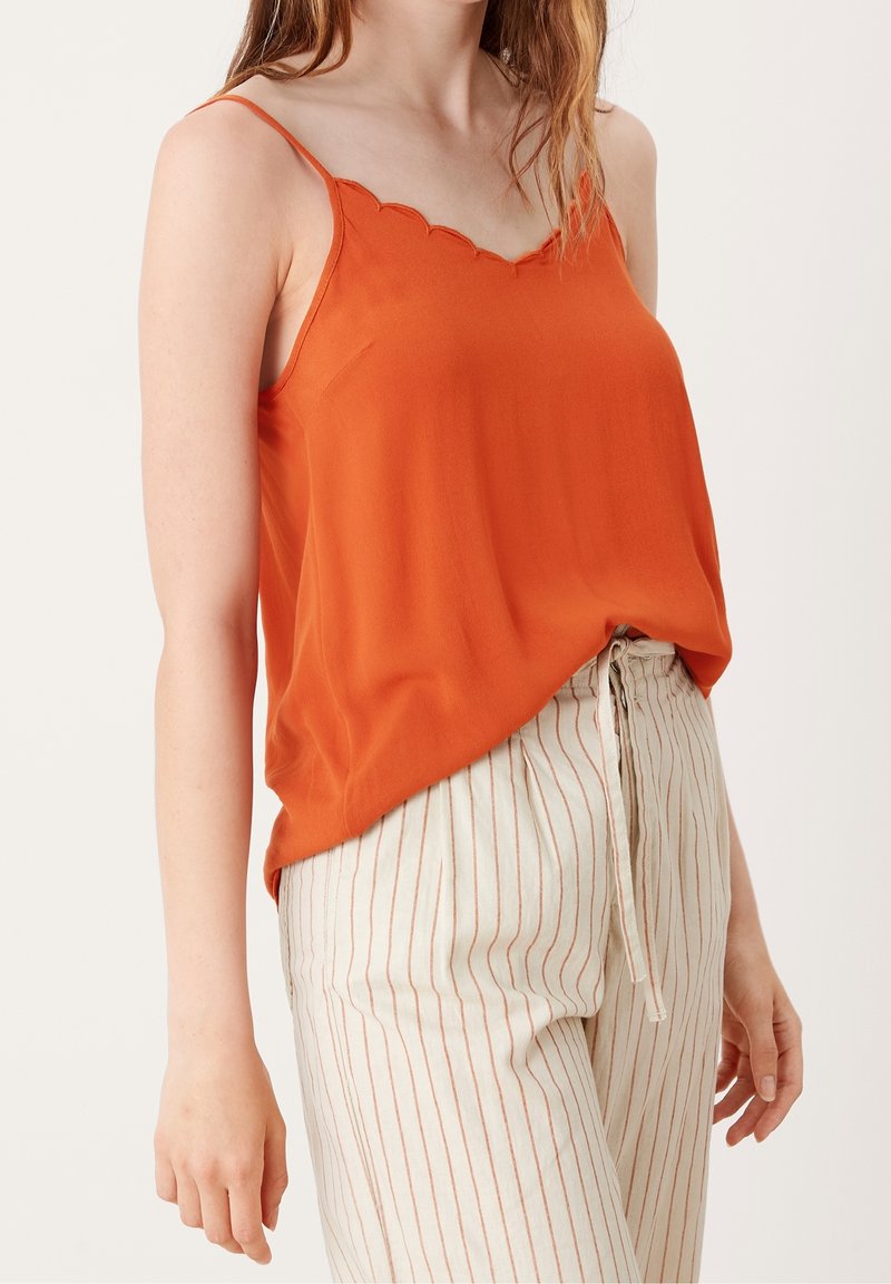 Femme portant un haut sans manches orange avec bord festonné et un pantalon crème taille haute à fines rayures verticales avec un nœud devant.