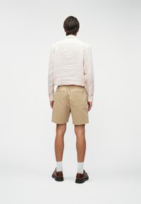 Chemise à rayures rose clair à manches longues, short beige, chaussures marron, chaussettes blanches, vue de derrière contre un fond uni.