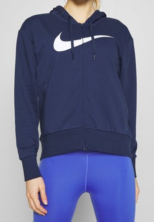 Kobieta w granatowej bluzie z kapturem zapinanej na zamek błyskawiczny z białym logo Nike i jaskrawo niebieskich legginsach, stojąca na jednolitym tle.