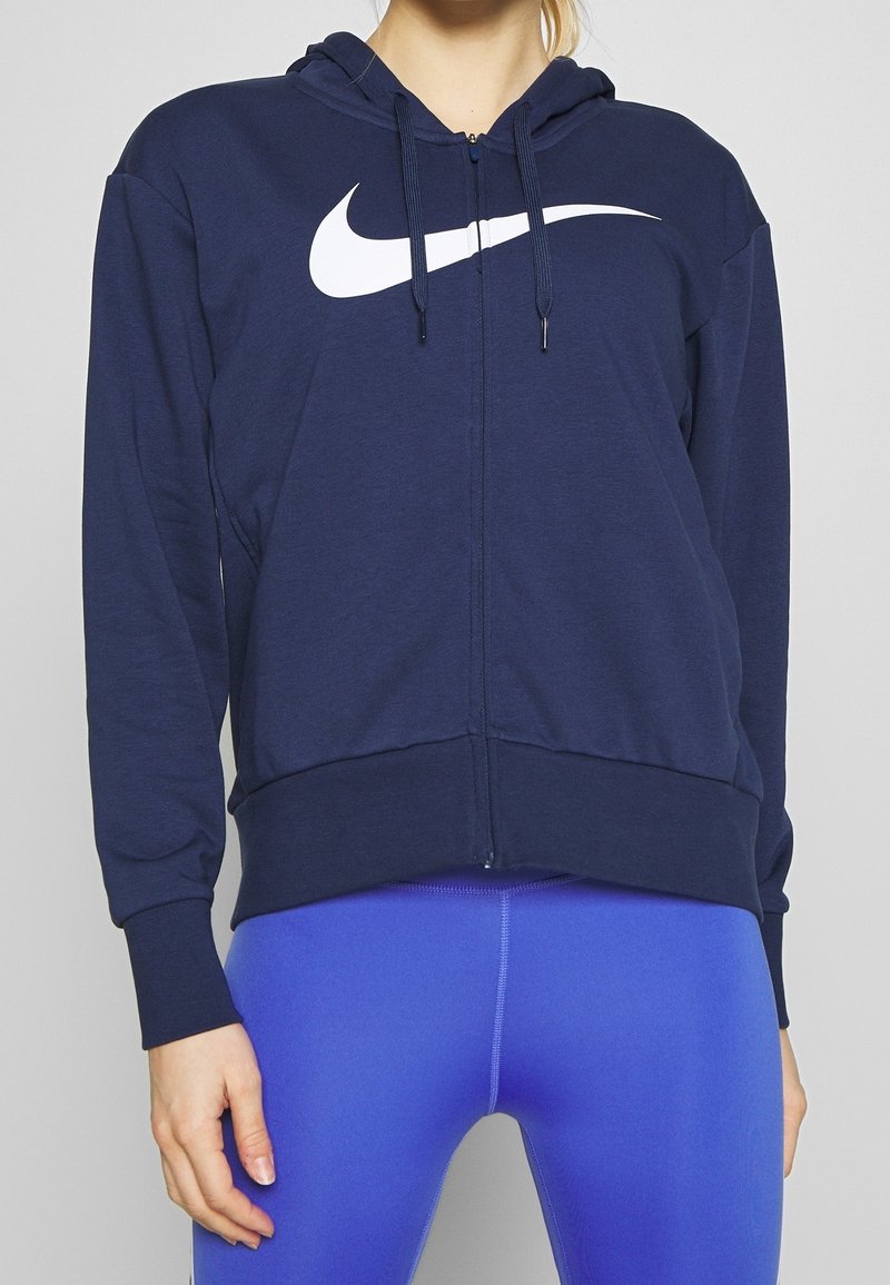 Femme portant un sweat à capuche zippé bleu marine avec logo Nike blanc et des leggings bleu vif, debout devant un fond uni.
