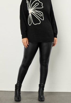 Czarny sweter z białym wzorem kwiatowym, w połączeniu z błyszczącymi czarnymi legginsami i botkami do kostki, na jasnym tle.