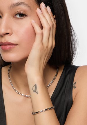 Silberne Gliederkette und Armband an einem Modell. Die Kette besteht aus quadratischen Gliedern; beide Stücke haben eine glatte Oberfläche und einen dezenten Glanz.
