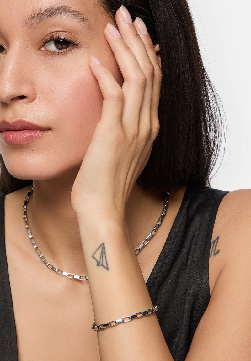 Sølv halskæde og armbånd på en model. Halskæden har firkantede led; begge stykker har en glat overflade og en subtil glans.