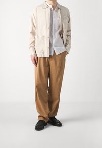 Chaqueta de algodón beige con botones en el frente y bolsillos, sobre una camisa blanca con cuello, combinada con pantalones marrones sueltos y zapatos negros.