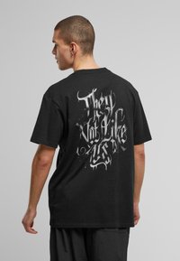 Black short-sleeve t-shirt features large white graffiti-style text on the back. Material appears soft with a relaxed fit and crew neck.