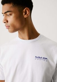Balts kokvilnas T-krekls ar zilu "Red Bull Racing" logo uz krūtīm. Klasiska apaļa kakla forma ar īsām piedurknēm.