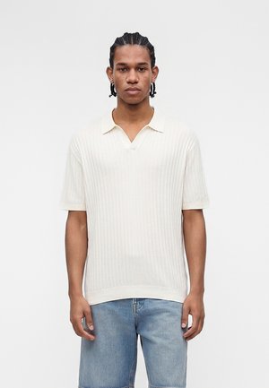 Joven con el pelo trenzado que lleva una camiseta polo blanca de manga corta y acanalada y pantalones cortos de mezclilla azul claro, de pie frente a un fondo blanco liso.