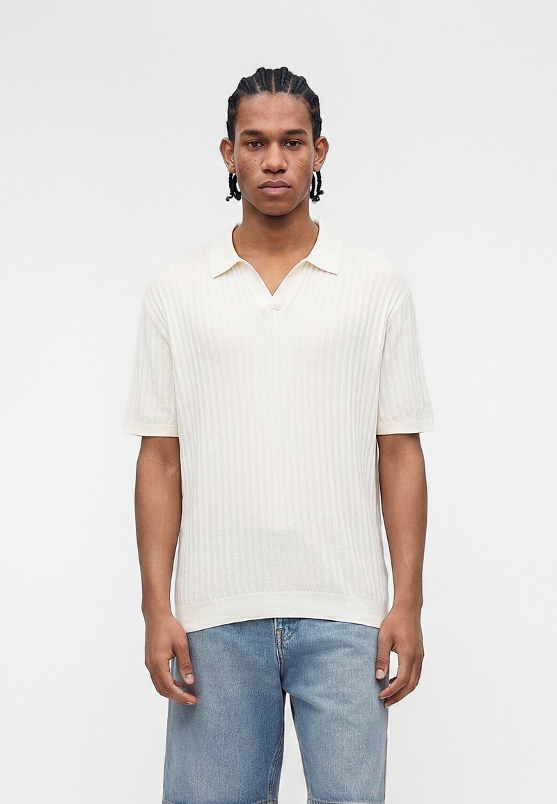 Jeune homme aux cheveux tressés, portant un polo blanc côtelé à manches courtes et un short en jean bleu clair, debout devant un fond blanc uni.