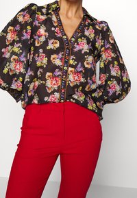 Blouse noire à fleurs avec des manches bouffantes, des motifs floraux colorés, et un devant à boutons avec un col et des détails de bordure contrastants.