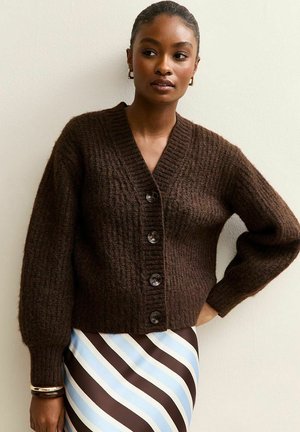 Bruine gebreide cardigan met een V-hals, zes knopen aan de voorkant en ballonmouwen, gedragen over een blauw-bruine gestreepte rok.