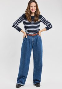 Stribet navy og hvid langærmet skjorte, blå brede jeans, brun bælte, sorte sko; model står med hænderne på hofterne.