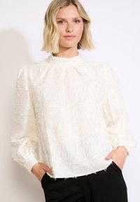 Blusa bianca con motivo floreale testurizzato, colletto alto e maniche a sbuffo, abbinata a pantaloni neri. Tessuto morbido e leggero.
